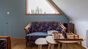 A room with a sloped ceiling, a sofa with floral upholstery, a wooden table with chess pieces, and a window with a view of trees. - 'Maison De Charlie' by Madame est Servie – private terrace, garden, and Wi-Fi (Trégastel)