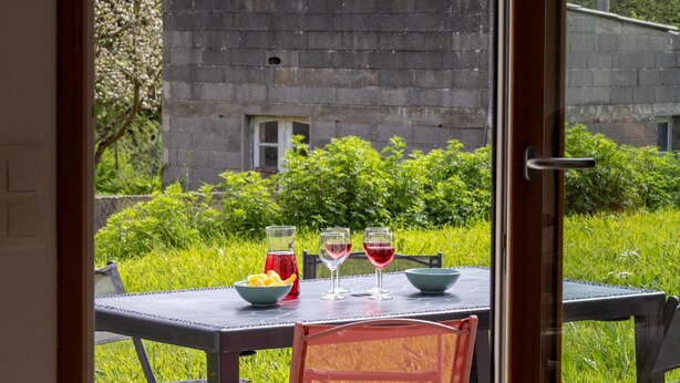 A dining table with a bowl of fruit, two wine glasses, and a pitcher, set against a backdrop of greenery and a stone building. - 'Maison De Charlie' by Madame est Servie – private terrace, garden, and Wi-Fi (Trégastel)