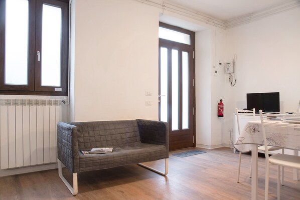 Une pièce avec un canapé gris, une table à manger avec des chaises, un téléviseur et une porte avec une fenêtre.