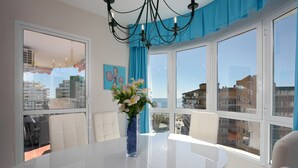 A modern room with a white table, a vase with flowers, a chandelier, and large windows with blue curtains.