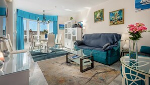 A modern living room with a glass coffee table, teal sofa, and a dining area with chairs and a table.
