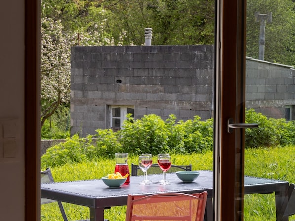 A dining table with a bowl of fruit, two wine glasses, and a pitcher, set against a backdrop of greenery and a stone building.