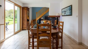 Un espace repas avec une table et des chaises en bois, un escalier, une cheminée et une grande fenêtre donnant sur un paysage verdoyant.