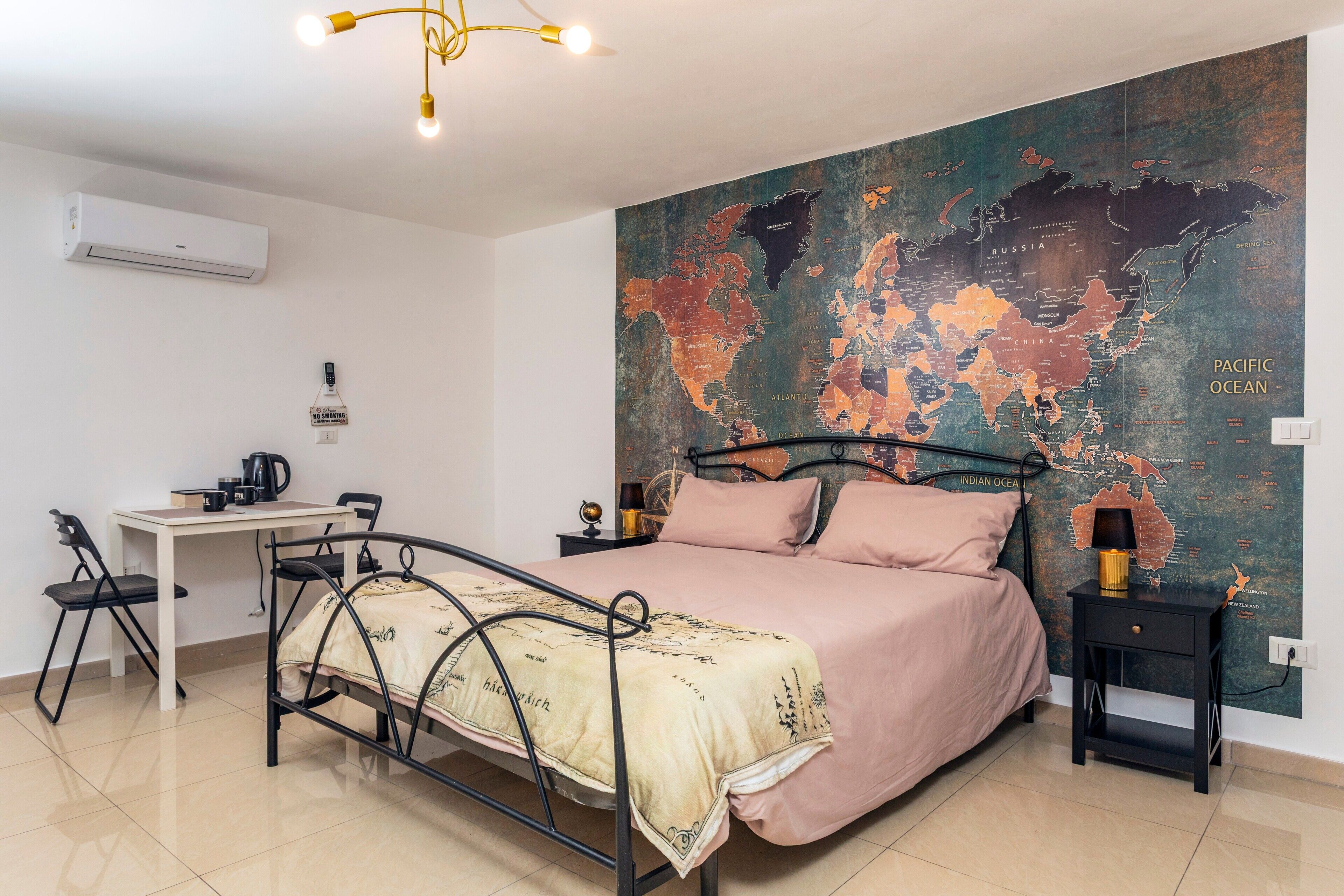 A bedroom with a metal bed frame, a desk, a chair, a small table with a lamp, and a world map mural on the wall.