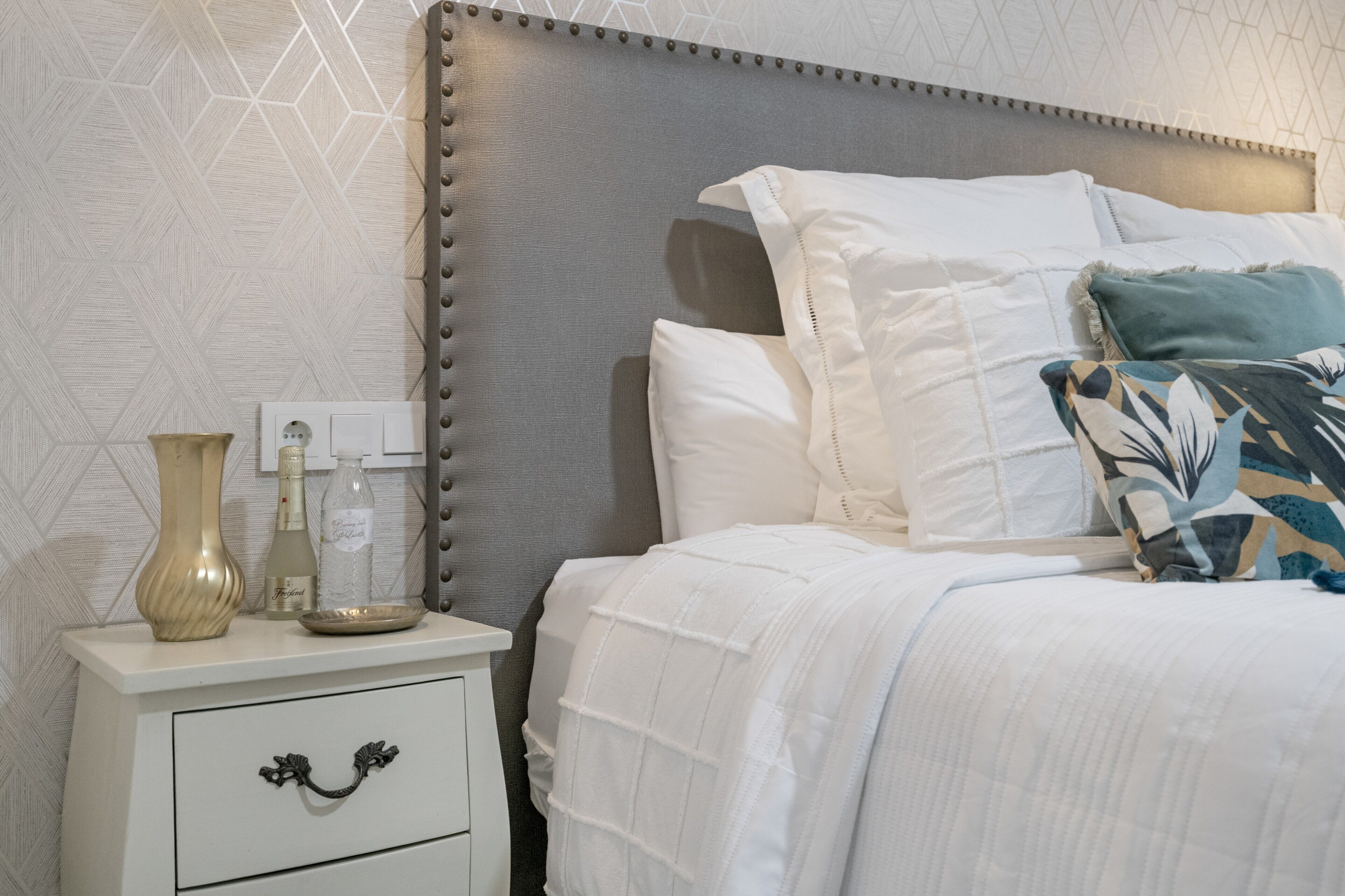 A neatly made bed with white bedding, a grey upholstered headboard, and decorative pillows.