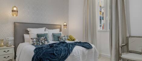 Un dormitorio con cama, mesitas de noche, una silla y un ventanal con cortinas.