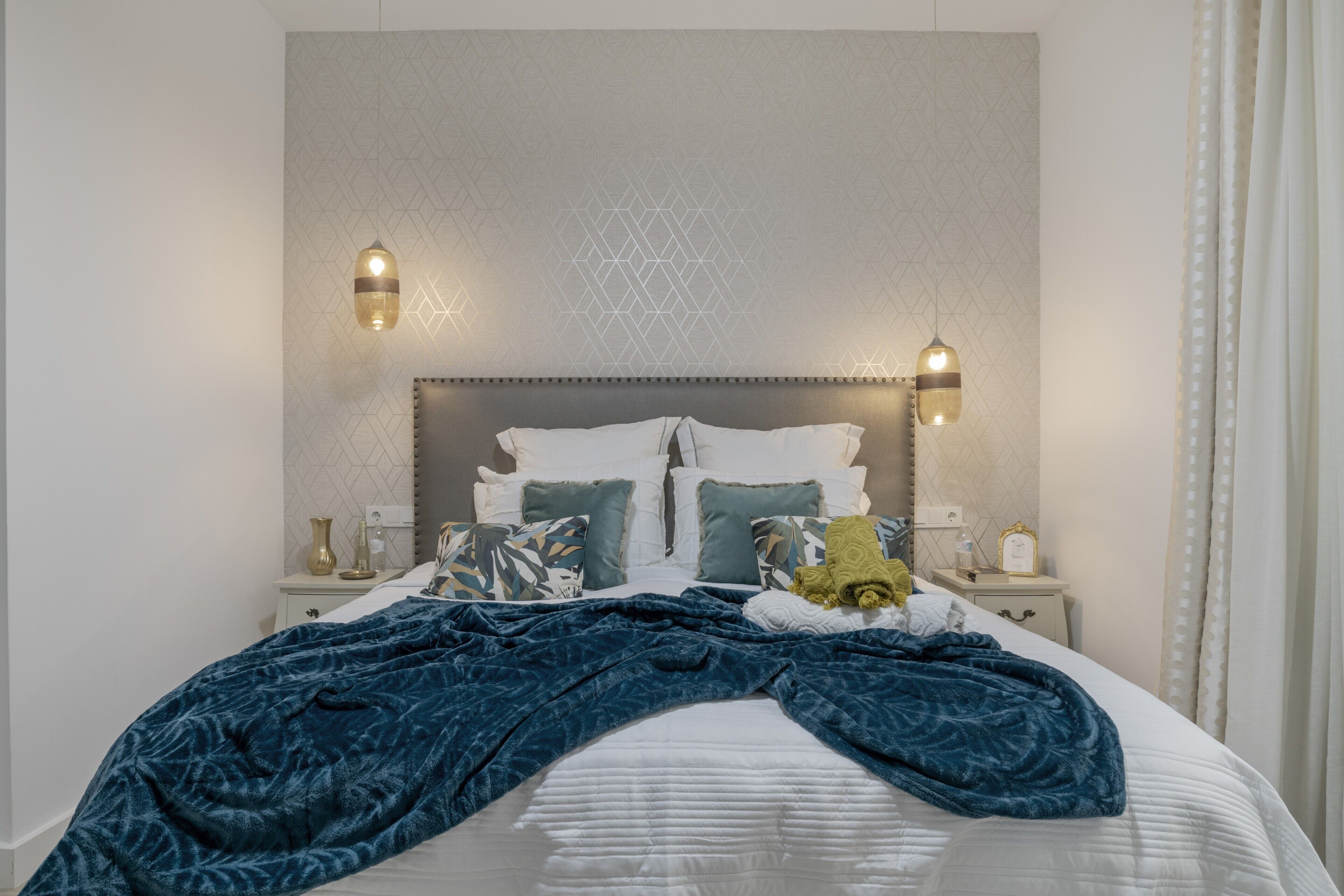 Una cama bien hecha con una colcha estampada, dos mesitas de noche con lámparas y una pared con diseño geométrico.