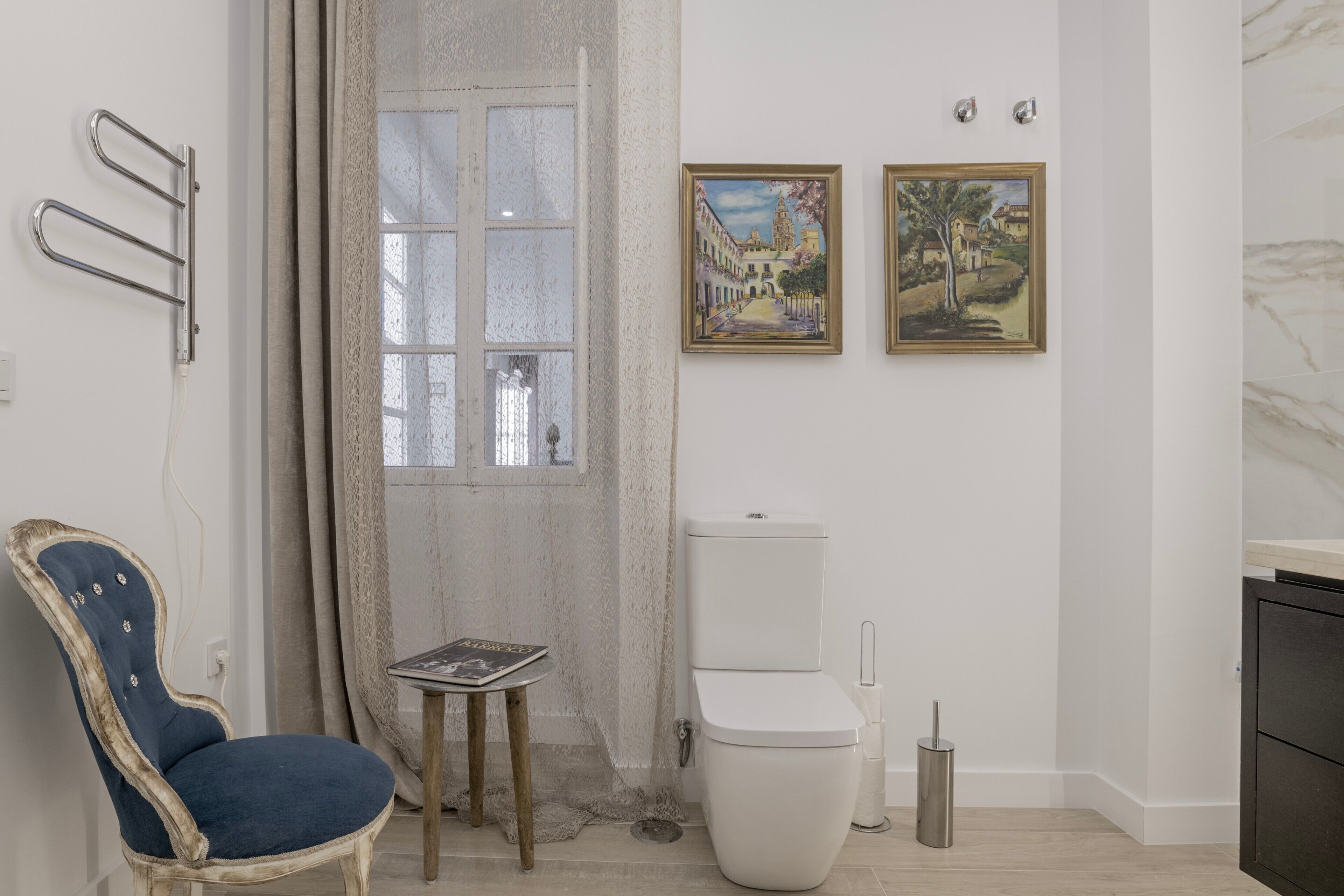 A bathroom with a toilet, a chair, a small table, and two framed pictures on the wall.