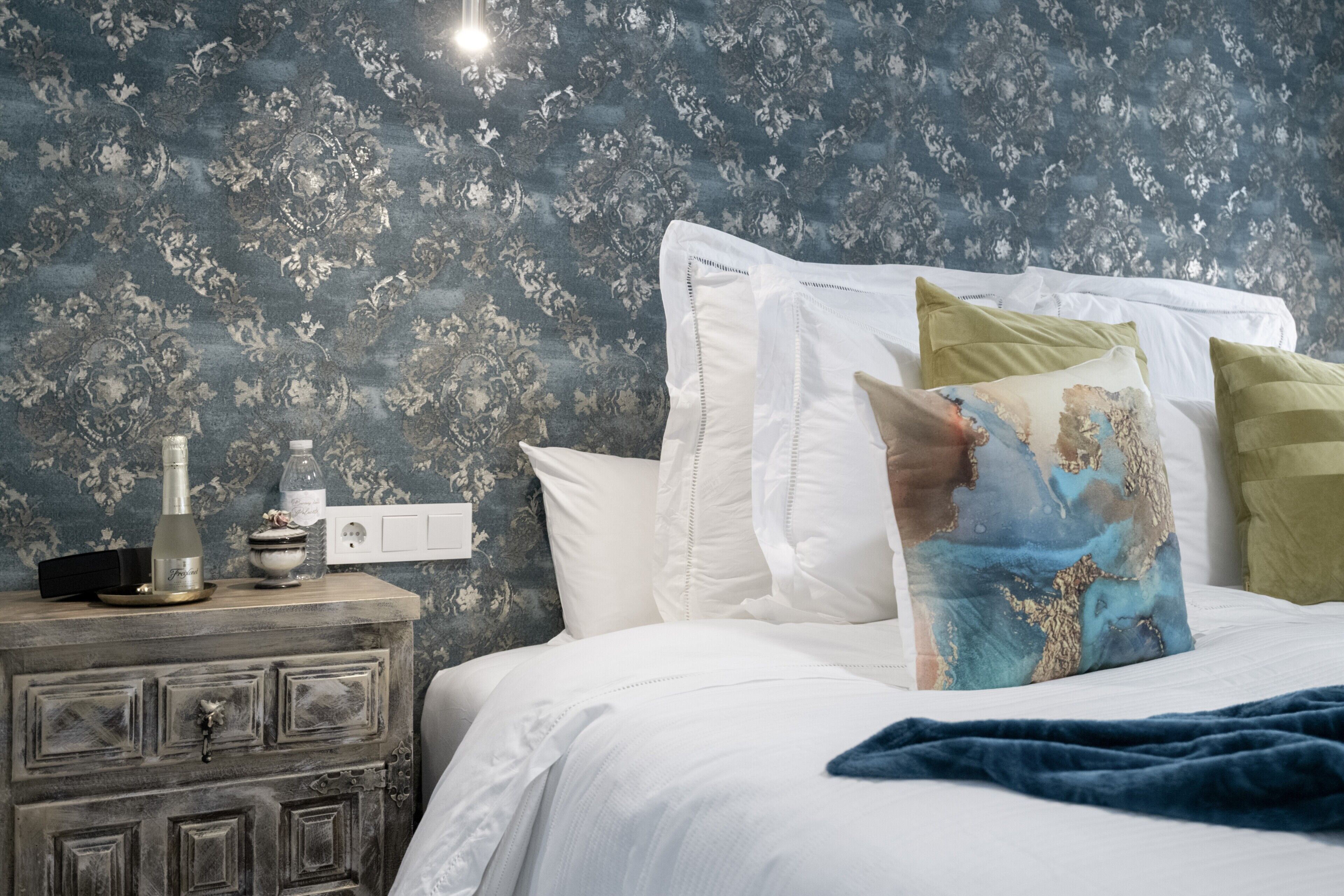 Un dormitorio con papel tapiz floral, una cama blanca con muchos almohadones, una mesita de noche con lámpara y una manta azul.