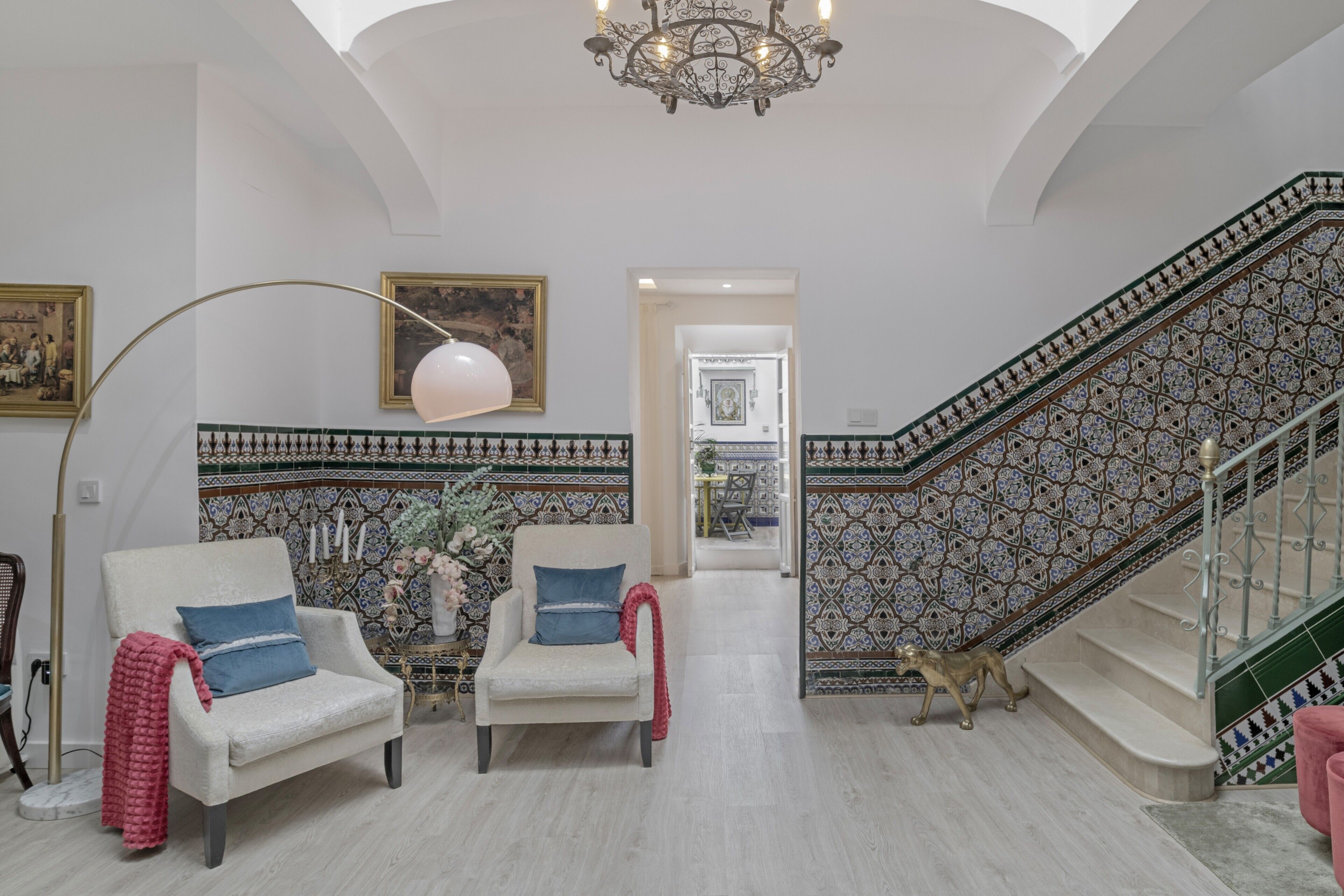 A spacious living area with two armchairs, a chandelier, a staircase with patterned tiles, and a hallway leading to another room.