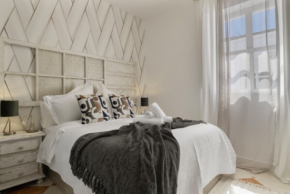 Un lit bien fait, avec une tête de lit blanche, un plaid gris et un coussin orné d’un motif géométrique.