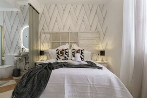 Un lit bien fait, avec une couette blanche et des oreillers, une table de chevet avec une lampe, un miroir et une salle de bain avec un lavabo et des toilettes blancs.