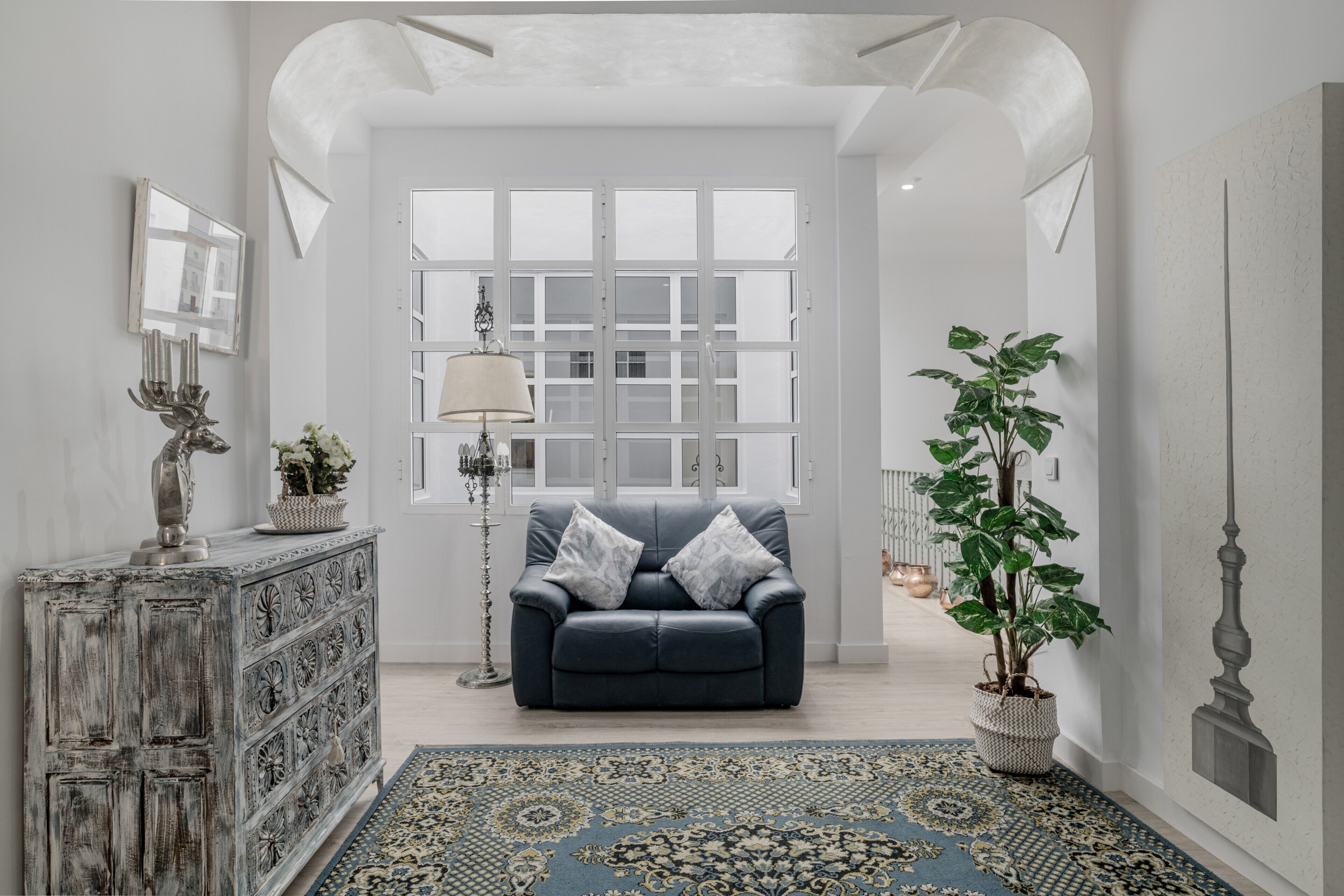 Una habitación con un sofá azul oscuro, un tapete estampado, un mueble blanco y una planta en maceta.