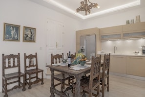 Uma sala de jantar com mesa de madeira, cadeiras e um lustre. Há uma geladeira e uma pia na área de cozinha adjacente.