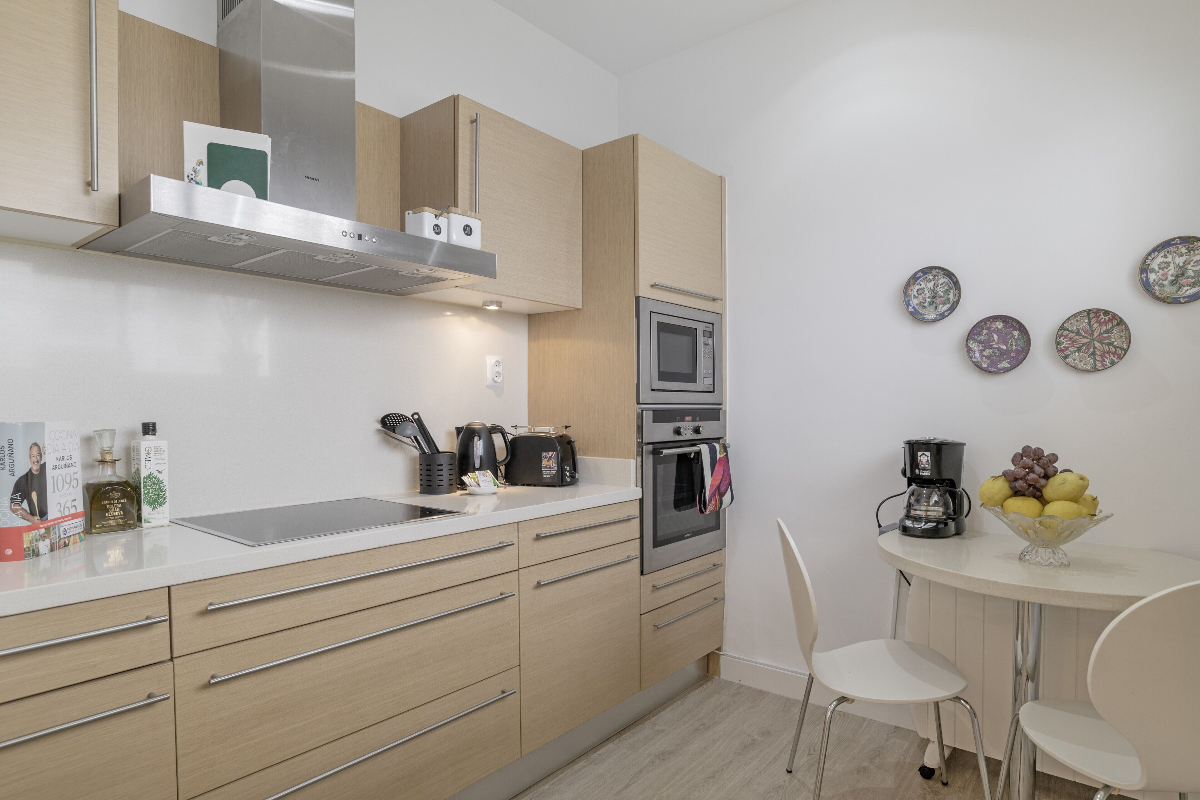 A modern kitchen with a microwave, oven, and coffee maker. There are white chairs and a small table with fruit.