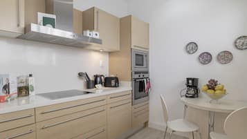 A modern kitchen with a microwave, oven, and coffee maker. There are white chairs and a small table with fruit.