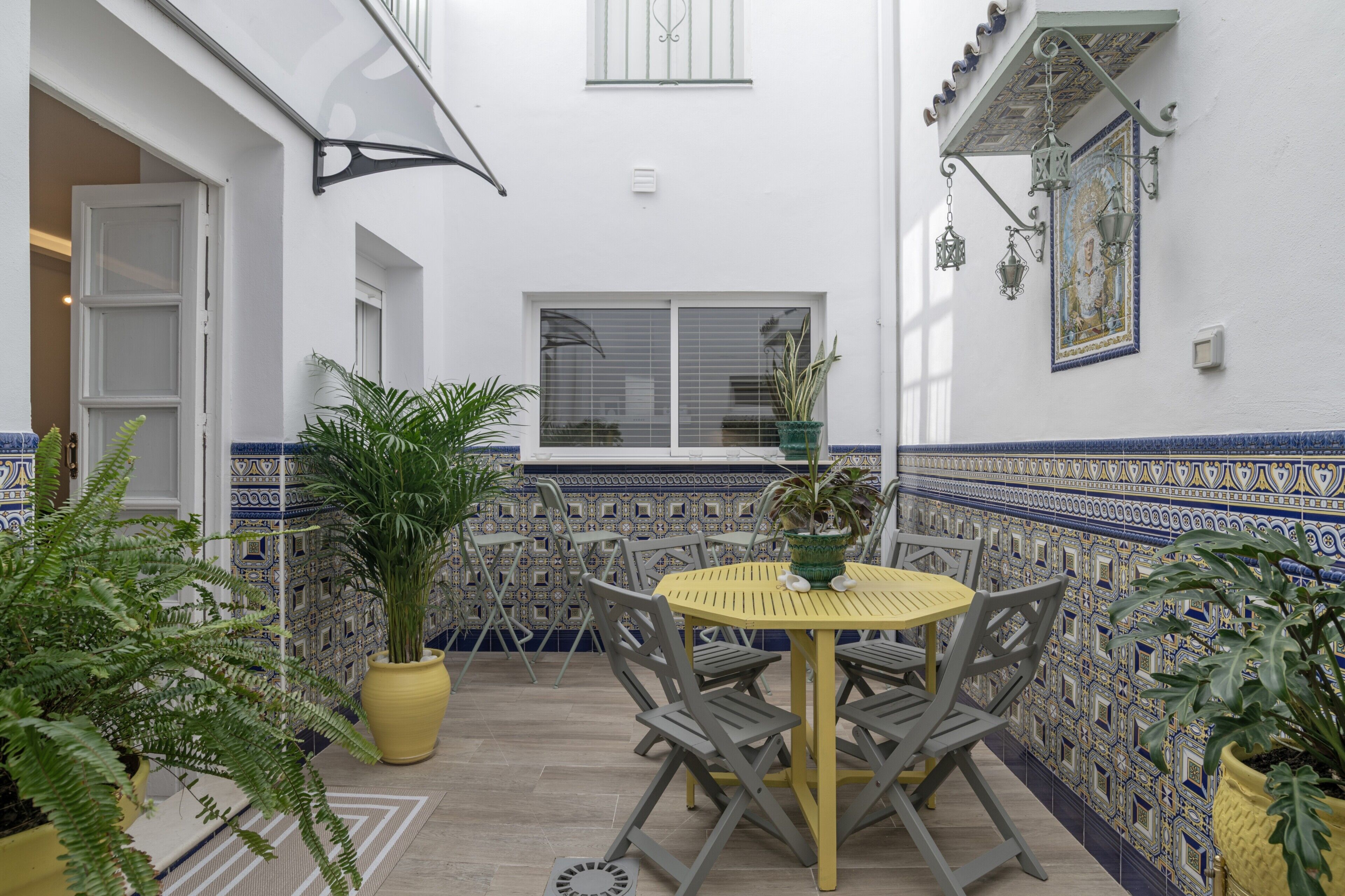Un patio con una mesa redonda y sillas, macetas con plantas y una pared con diseño geométrico de azulejos.