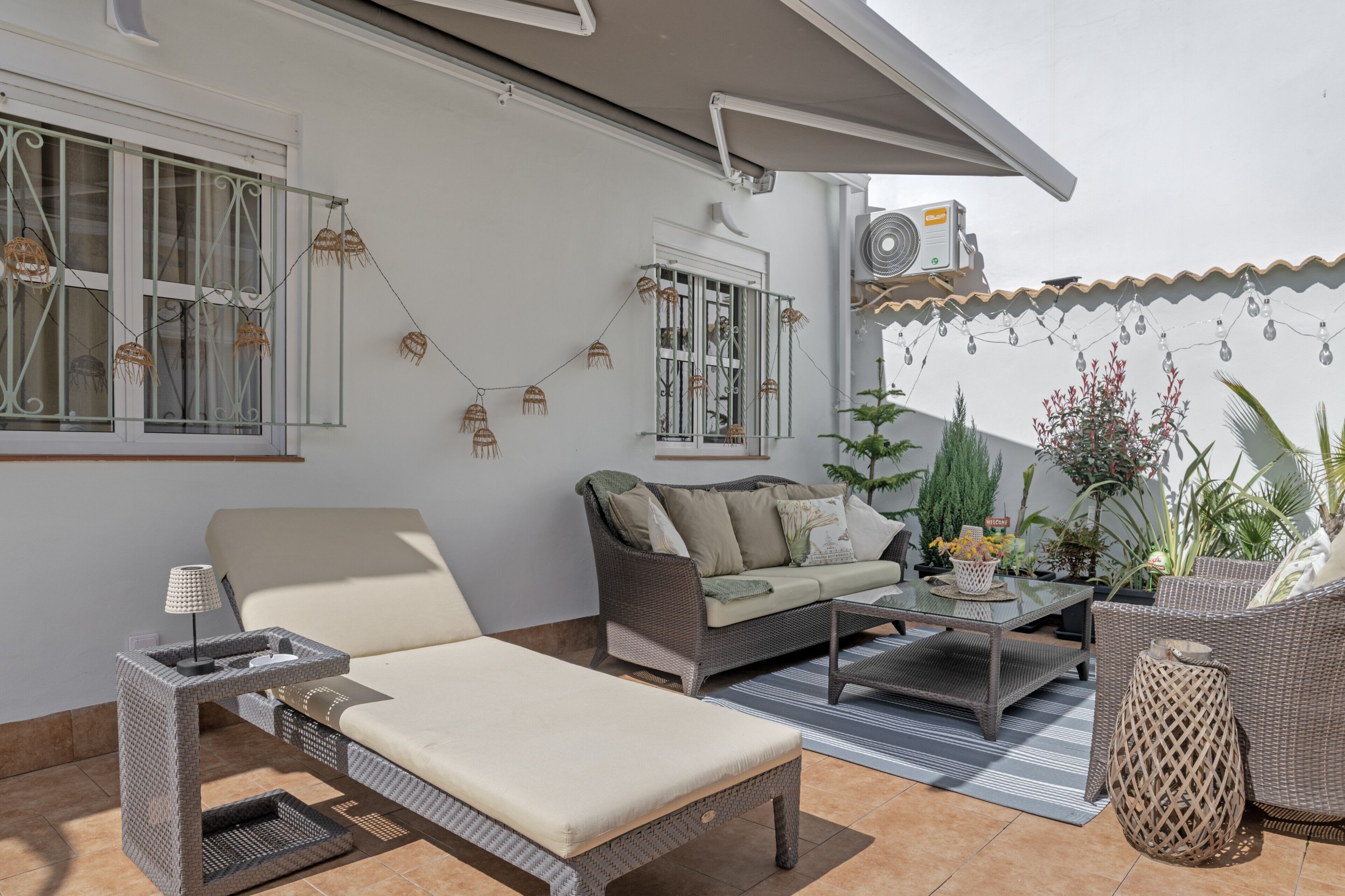 A patio with a sofa, lounge chair, and table, surrounded by potted plants and decorative lights.