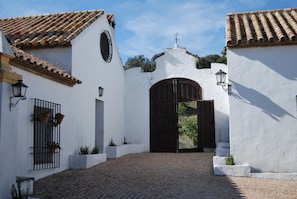 Un patio con un sendero adoquinado, un gran portón de madera y edificios blancos con techos de tejas.