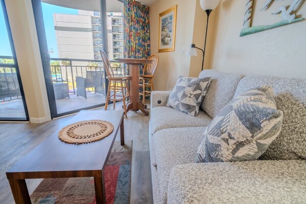 A living room with a sofa, a coffee table, and a chair. There is a balcony with a view of buildings. - Meridian 307 (Myrtle Beach)
