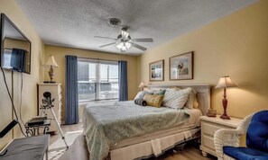 A hotel room with a bed, a ceiling fan, a desk with a lamp, and a chair.