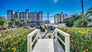 A wooden staircase leading to a building with multiple floors, surrounded by greenery and parked cars.