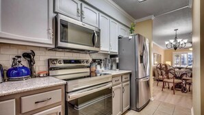 A kitchen with a microwave, stove, oven, and refrigerator. There is a dining area with chairs and a chandelier.