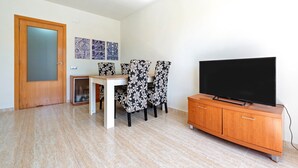 A living room with a TV, a wooden cabinet, a dining table, and four patterned chairs.