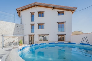 Un edificio de dos pisos con piscina, mobiliario exterior y un muro de piedra.
