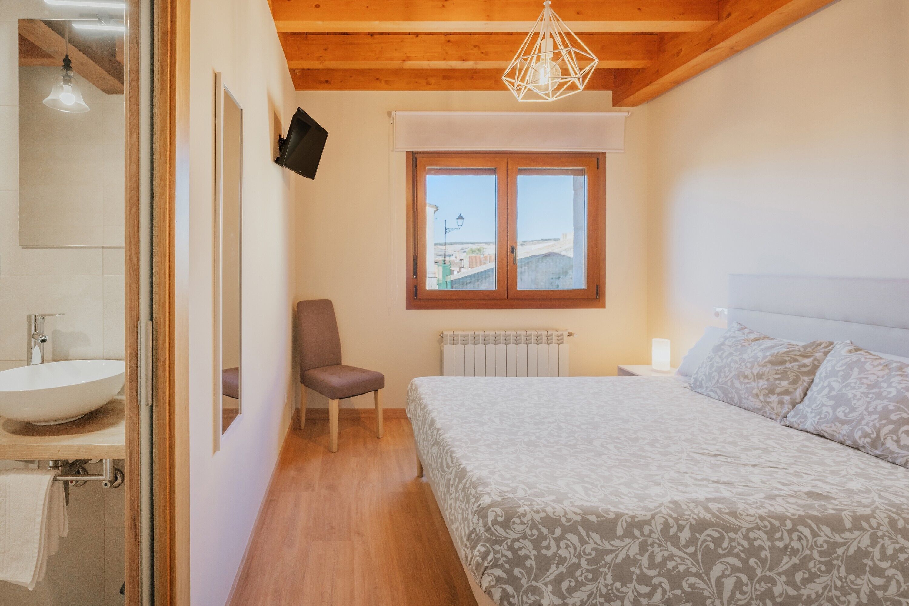 A bedroom with a bed, a chair, a window with a view, a radiator, and a bathroom with a sink and mirror.