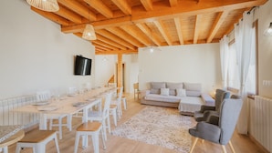 A dining area with a long table and chairs, a sofa, and a television on the wall.