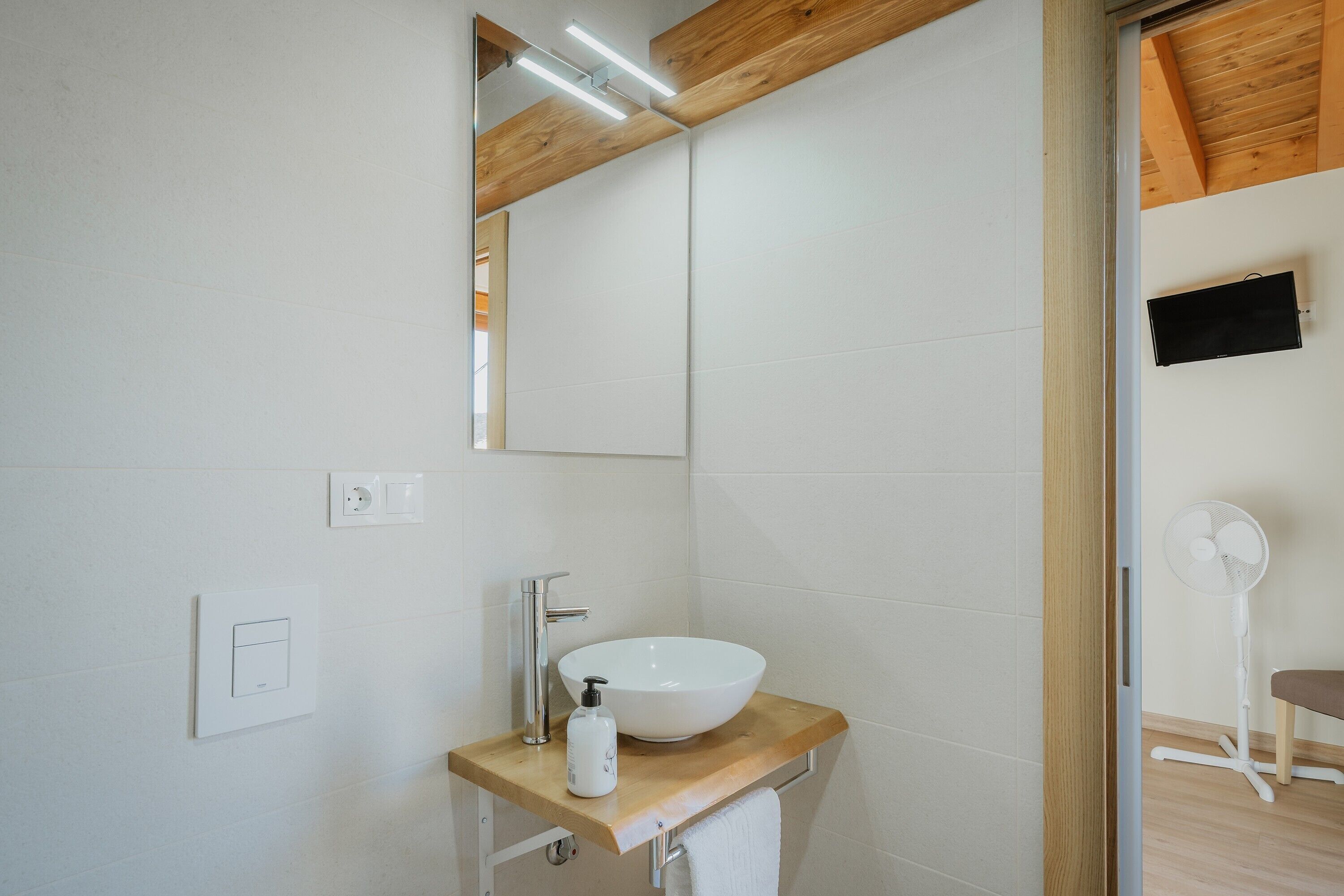 Bagno con piano in legno, lavandino bianco, specchio, ventola a parete e interruttore della luce visibile.