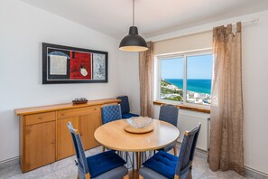 Ein Essbereich mit einem runden Tisch, blauen Stühlen, einem Holzschrank und Blick auf das Meer.