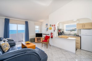 Un espace de vie comprenant un canapé, un téléviseur, une table à manger, une cuisine équipée d’un réfrigérateur, et une vue sur la mer accessible par une porte coulissante.