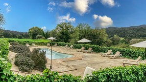 A pool area with lounge chairs and umbrellas, surrounded by greenery and hills.