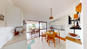A compact kitchen with white cabinets, a dining area with wooden chairs, and a view through a glass door. - Villa 'Ersilia' with Sea View, Wi-Fi and Air Conditioning (Campulongu)