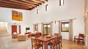A spacious living area with a wooden dining table, chairs, a sofa, and a staircase leading to an upper level. - Country House 'Sant Salvador' with Mountain View, Wi-Fi and Air Conditioning (Illes Balears)