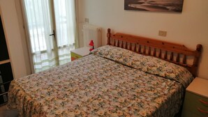 A bedroom with a bed, a nightstand, a lamp, a radiator, and a painting on the wall. - Euroholiday Village 2A Apartment with Private Terrace, Shared Garden and Wi-Fi (Jesolo)