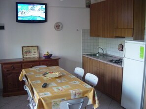 Une petite cuisine équipée d’une table à manger, de chaises, d’un réfrigérateur, d’un évier et d’un téléviseur fixé au mur.