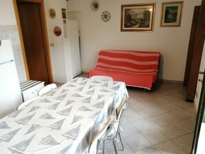 Une petite pièce propre et bien rangée, avec un lit, une table et une chaise. Il y a deux tableaux encadrés au mur, un réfrigérateur et un placard.