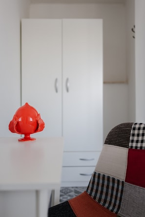 Un meuble blanc à deux portes, un objet rouge posé sur une surface blanche et un coussin à carreaux.