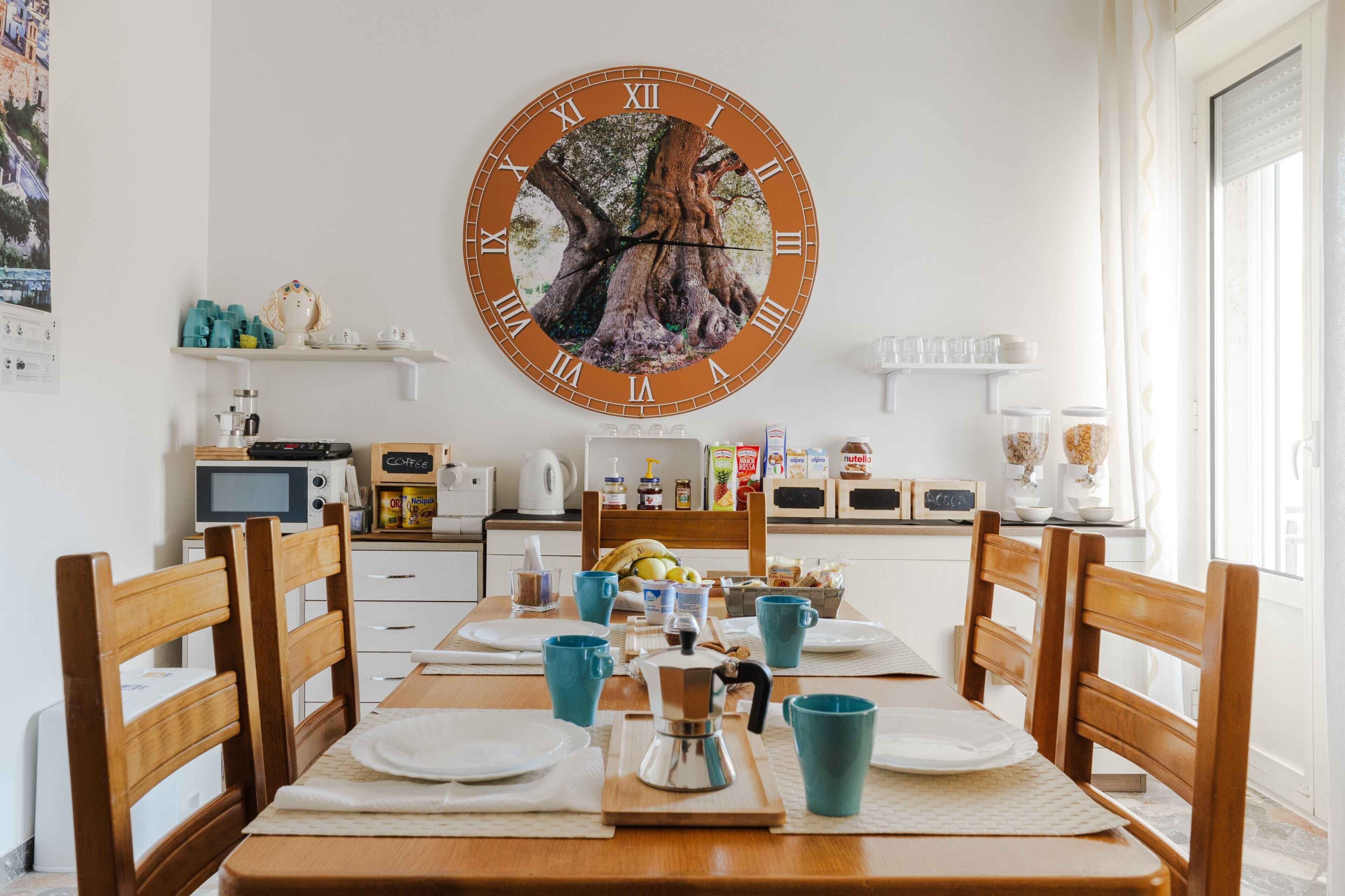 A dining area with a wooden table, chairs, a large wall clock, and kitchen appliances in the background.