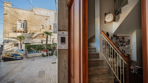 A stone building with a sign reading 'PLAZZA N'EGNA,' a staircase with a wooden railing, and a wall with a clock.