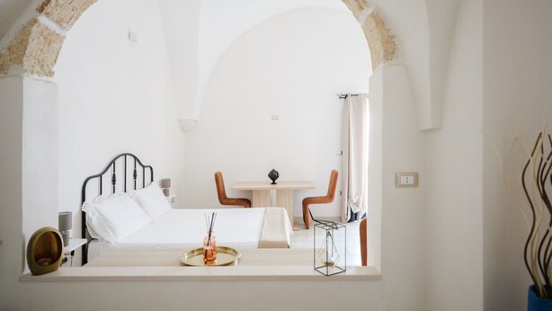 A room with a bed, a table, and chairs, an arched doorway, and a vase with branches.