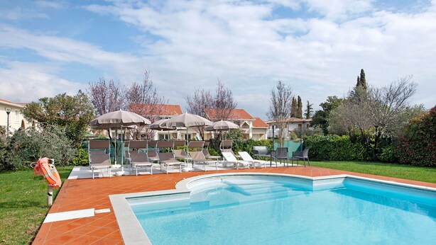 Une piscine entourée d’un jardin et de bâtiments, avec des chaises longues et des parasols.
