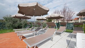 Une rangée de chaises longues dotées de coussins beiges et de structures blanches, disposées sous des parasols beiges sur une terrasse carrelée.