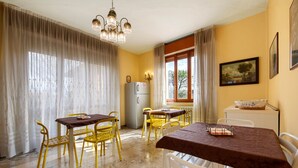 Un coin repas avec des chaises jaunes, une table recouverte d’une nappe marron, un lustre, un réfrigérateur et une fenêtre avec des rideaux.