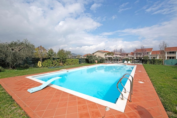 Una piscina rectangular con solárium empedrado, rodeada de vegetación y edificios al fondo.
