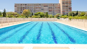 A swimming pool with a clear blue water surface, surrounded by a fence. In the background, there is a multi-story building with balconies and a garden area with various plants.