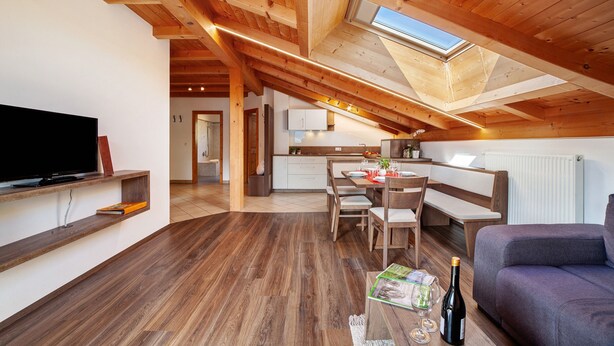 A modern loft-style room with a wooden ceiling, a dining area, a kitchen, and a living room with a flat-screen TV and seating.